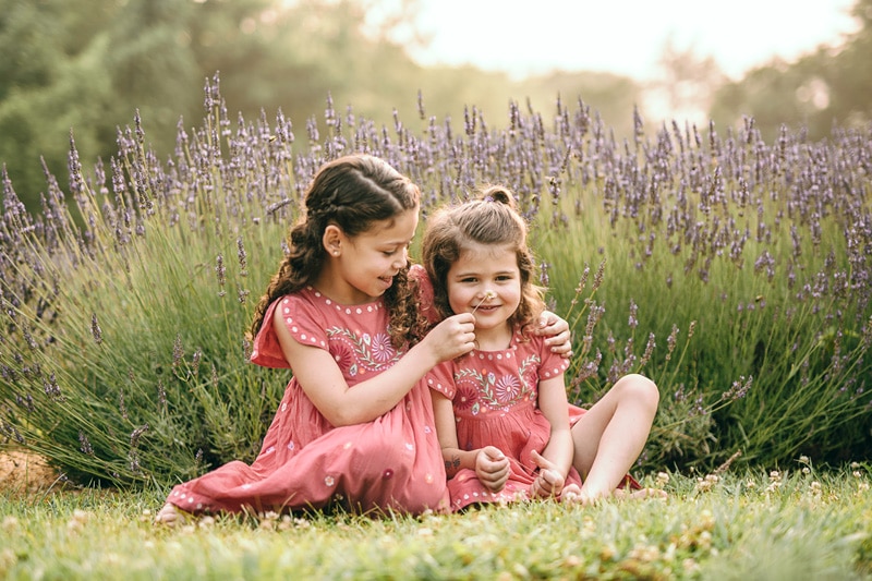 an older sister in red dress is tickling a nose of her baby sister with a flower at the lavender farm during a summer family session by Bella Luna Photography in Loudoun County