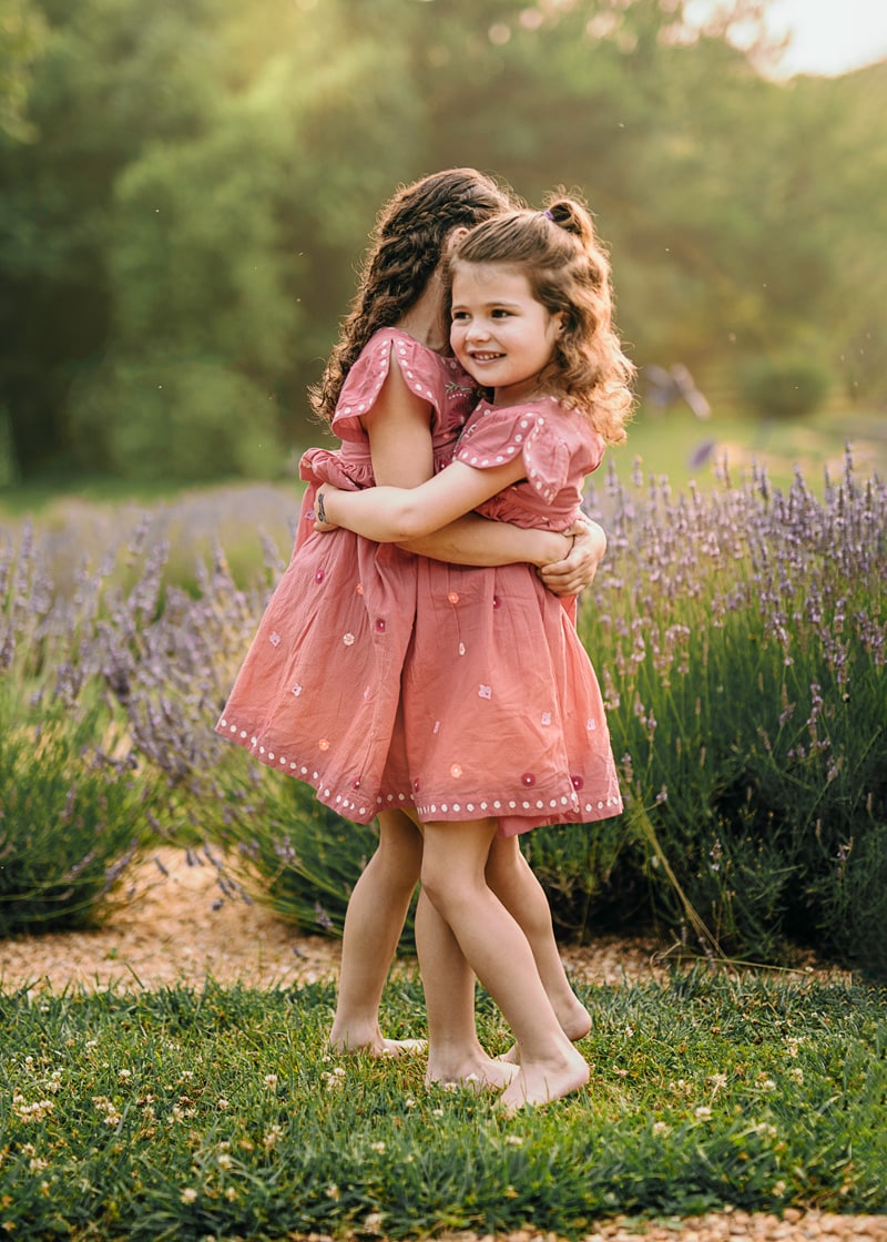 sisters in red dresses hugging next to lavender during an outside summer family session in Loudoun County by Bella Luna Photography