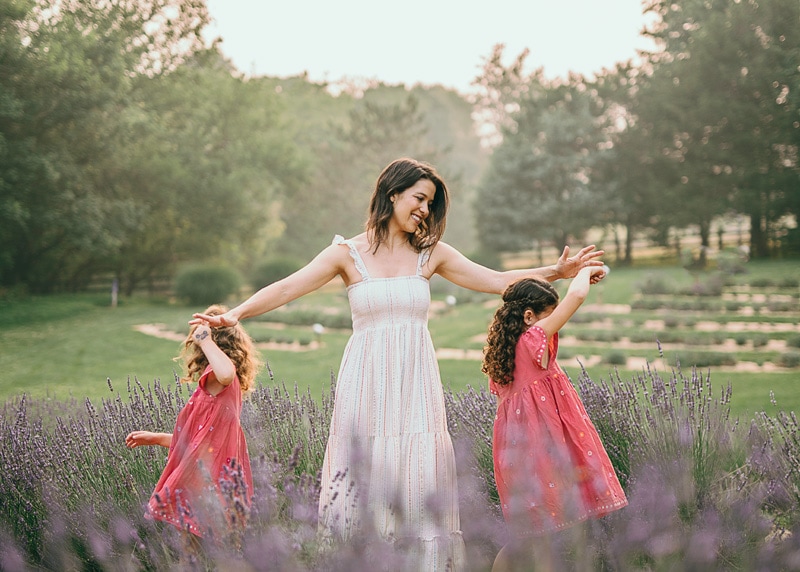 A mom in white dress twirling with her daughters at lavender farm during a summer outside family session by Bella Luna Photography in Leesburg, VA
