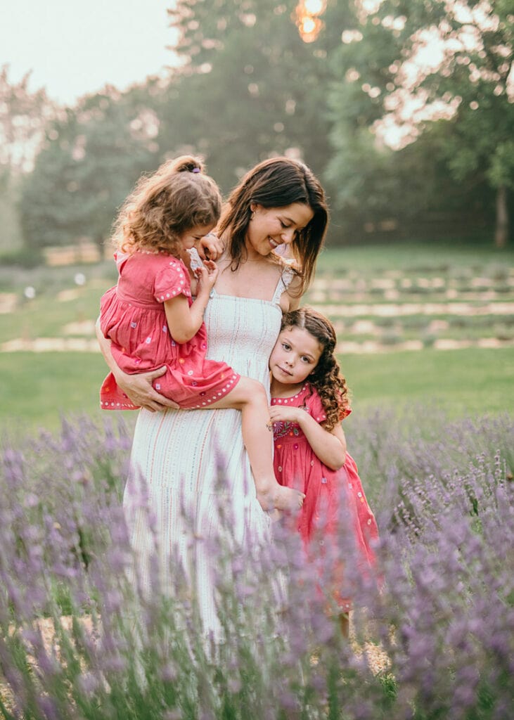 A mom is holding her younger daughter while looking down at her older daughter at the lavender farm during a summer family session by Bella Luna Photography in Leesburg, VA